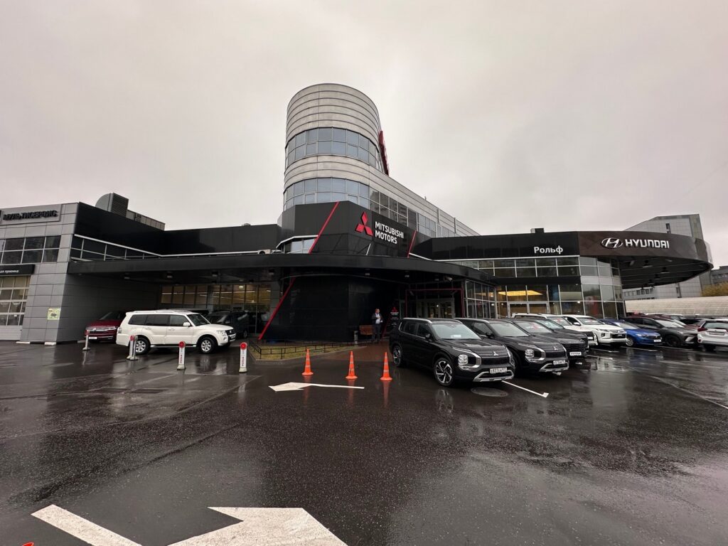 Фотография здания автосалона 'Рольф', представляющего марки Mitsubishi и Hyundai. Современное здание с цилиндрической башней и вывесками с логотипами брендов. На переднем плане - ряд автомобилей, выставленных на продажу.
