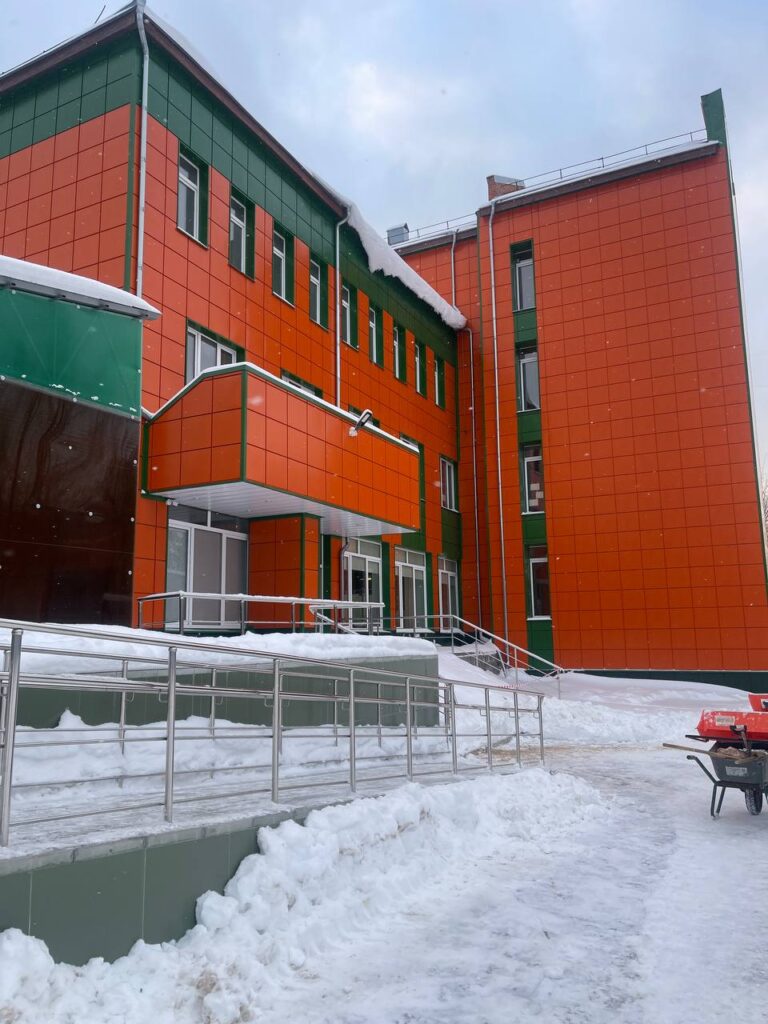 Фото здания с фасадом из оранжевых и зеленых панелей, крыльцом с пандусом и заснеженной территорией. Зимний вид.