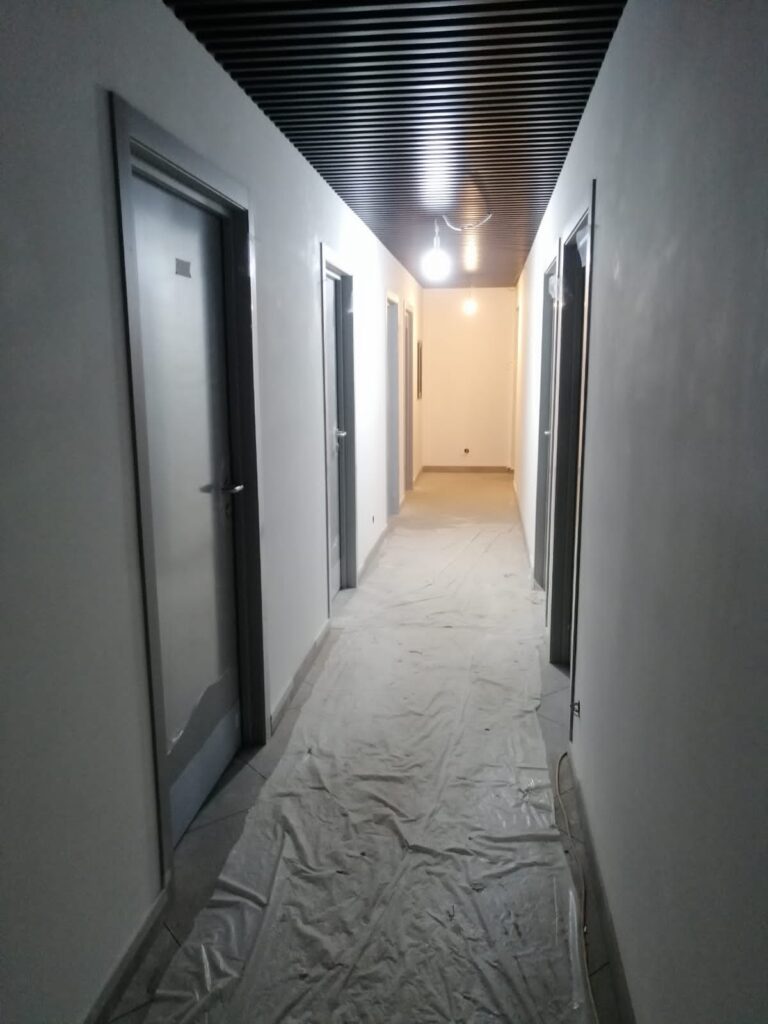 Длинный коридор с серыми дверями, чёрный реечный потолок, пол под защитной плёнкой, подвесные лампы, белые стены.