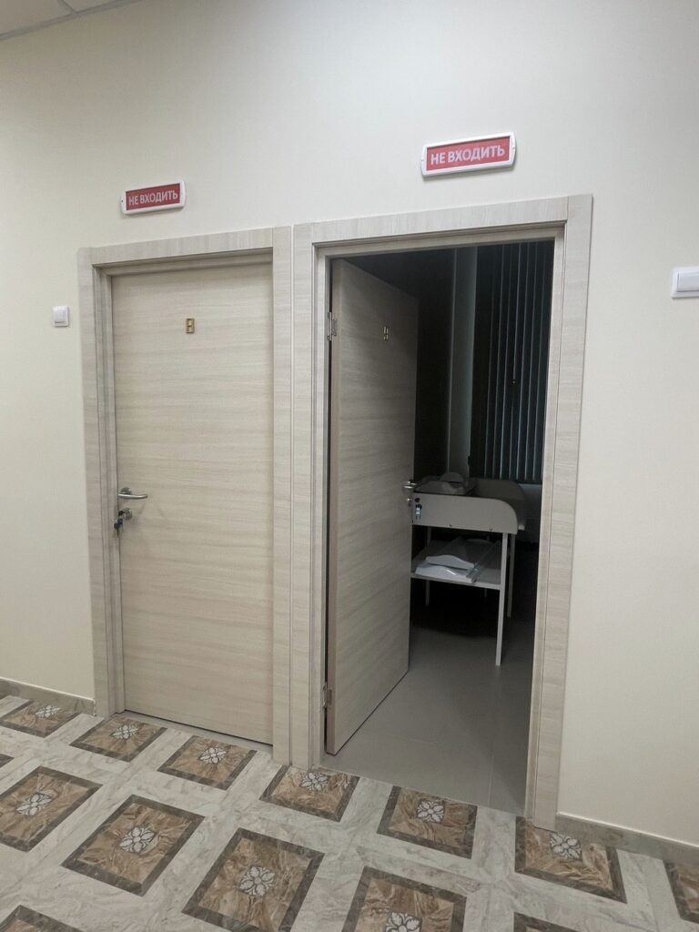 Две светлые двери с табличками «НЕ ВХОДИТЬ», одна открыта — вид на комнату со столом и жалюзи, узорчатый плиточный пол.