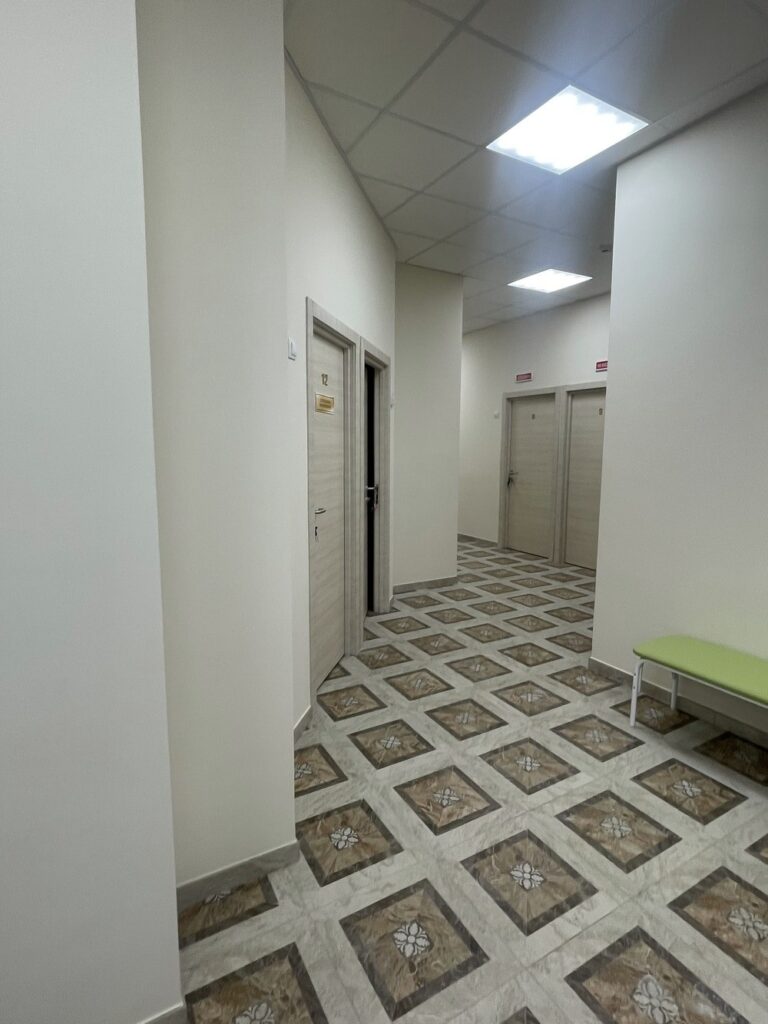 Коридор со светлыми дверями (номера 12 и 9), узорчатый плиточный пол, зелёная скамья справа, потолочные светильники.