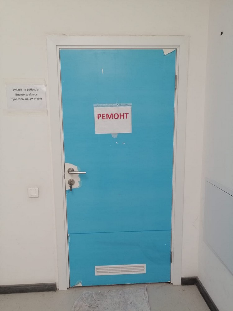 Голубая дверь с табличкой «РЕМОНТ», белая стена, серый пол, рядом — информационная записка о неработающем туалете.