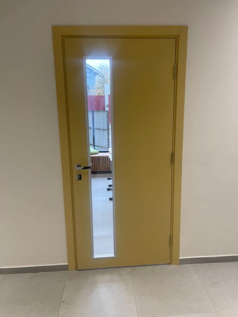 Желтая дверь с узким вертикальным стеклом и серебристой ручкой в помещении со светлыми стенами и плиточным полом