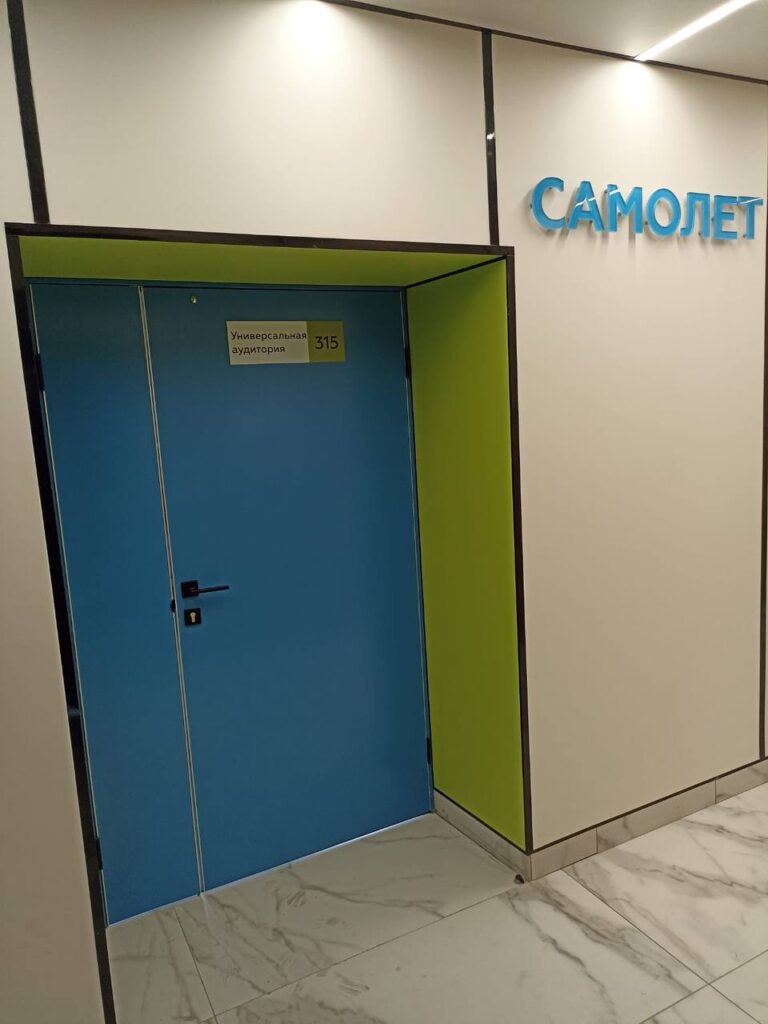 Синяя двустворчатая дверь с табличкой «Универсальная аудитория 315» в зелёном портале, рядом — надпись «САМОЛЕТ», мраморный пол.