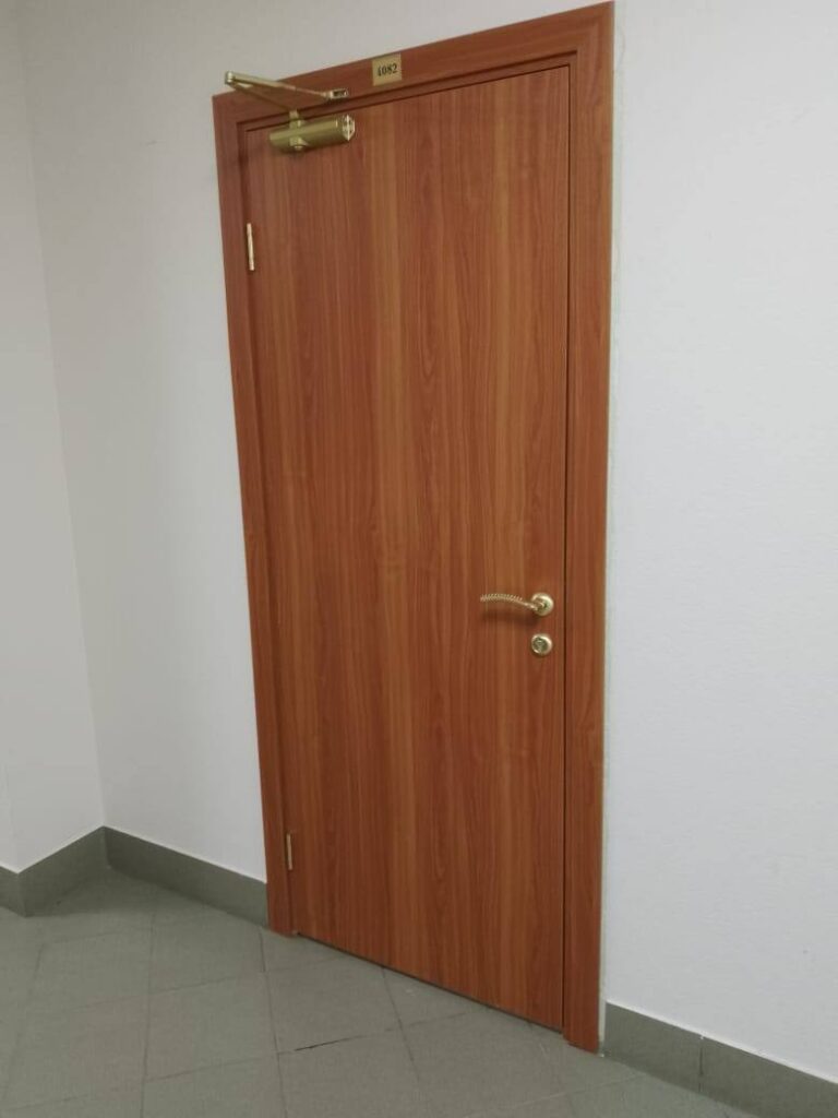 Дверь с деревянным шпоном, номер 4082, золотистая ручка и доводчик. Угол коридора, белые стены, серый пол.