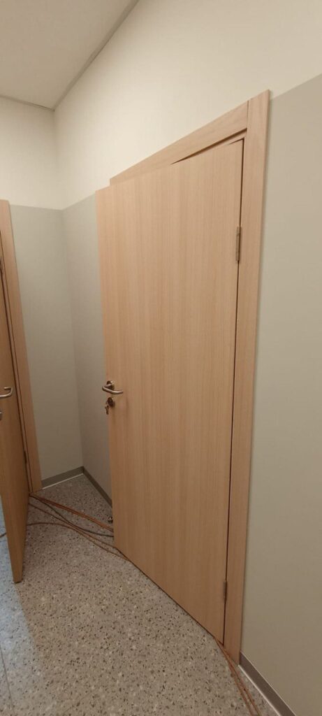 Две светлые двери под дерево в углу, правая приоткрыта с ключом в замке. На полу оранжевые провода, стены двухцветные.