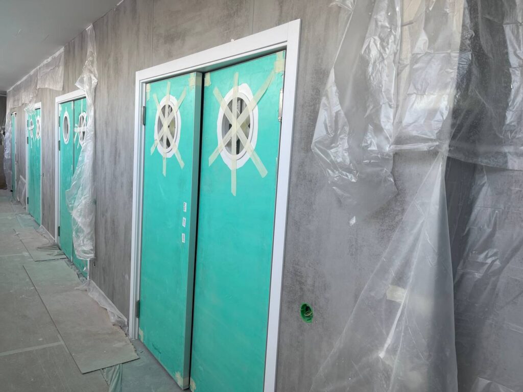 Двустворчатые двери в бирюзовой плёнке с круглыми окнами, заклеенными скотчем, на фоне белых стен и бетонного пола.