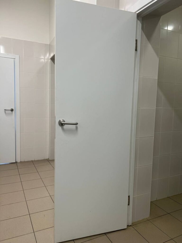 Белая дверь с металлической ручкой в помещении, отделанном светлой плиткой. На заднем плане видна ещё одна дверь.