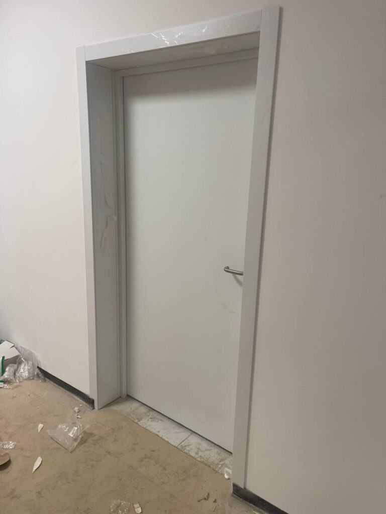 Белая дверь с ручкой в проёме, часть коробки ещё в плёнке. Пол не отделан, видны остатки упаковки у стены