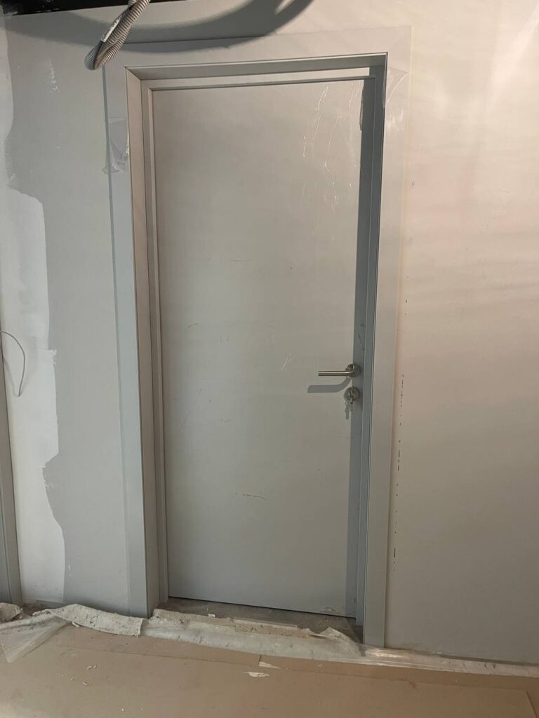Белая дверь с ручкой в светло-серой коробке на фоне стен со шпаклёвкой и гофротрубой сверху