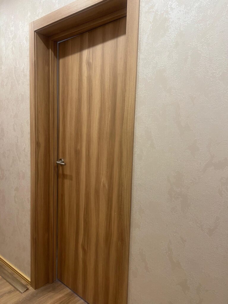Дверь цвета светлого дерева с широкой коробкой на фоне бежевой текстурированной стены.